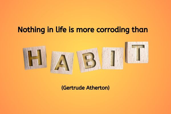 An ombre background of a yellow focal point in the centre of the image, radiating out to an orange on the outer edges of the image. The text reads: "Nothing in life is more corroding than habit." (Gertrude Atherton) The word habit is made up of wooden blocks, with one letter of the word carved into the top of the block.