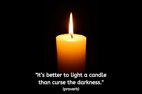 A black background has a single lit candle in the centre of the image, with the flame giving out an orange glow of light. The text reads: "It’s better to light a candle than curse the darkness." (Proverb)