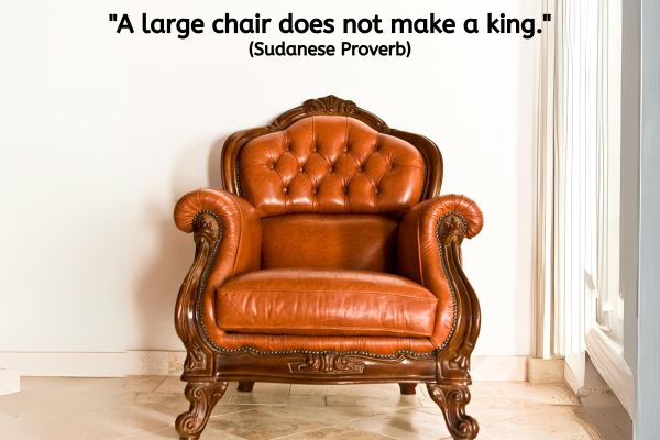 The image prominently features a luxurious and ornate armchair, crafted from rich brown leather with a deep tufted design on the backrest. The chair's arms and legs are adorned with intricate carvings, enhancing its regal and antique appearance. The leather itself is a vibrant, polished chestnut colour, indicating well-maintained quality and a stately presence. The chair sits against a plain, light-coloured background that accentuates its detailed craftsmanship and stately design. Above the chair, there is a Sudanese proverb: "A large chair does not make a king."