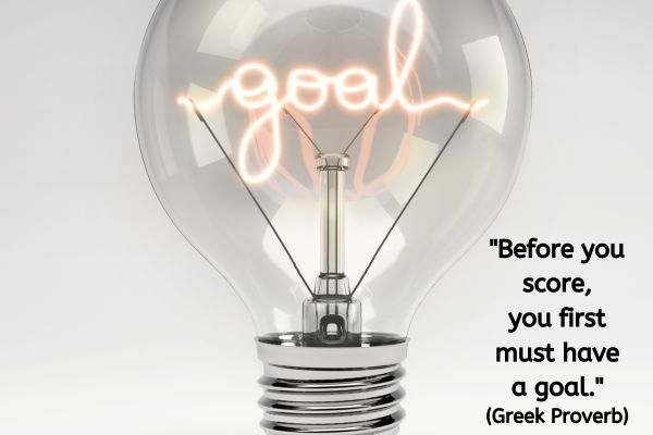 The image features a clear, large lightbulb against a plain, light gray background. Inside the lightbulb, the filament is uniquely shaped to spell out the word "goal" in a cursive, glowing neon style. Below the lightbulb, there is a Greek proverb: "Before you score, you first must have a goal."