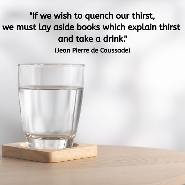 A clear glass of water sits on a wooden table, resting on a square wooden coaster. The background is softly lit and minimal. Overlaid text reads: “If we wish to quench our thirst, we must lay aside books which explain thirst and take a drink.” (Jean Pierre de Caussade)