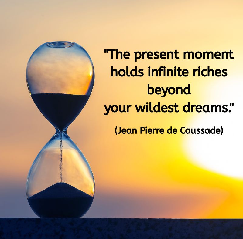 An hourglass stands against a golden sunset sky, the sand flowing steadily from top to bottom. Above it, in black text, is the quote: “The present moment holds infinite riches beyond your wildest dreams.” (Jean Pierre de Caussade) The image evokes stillness, time, and the quiet abundance of the present moment.