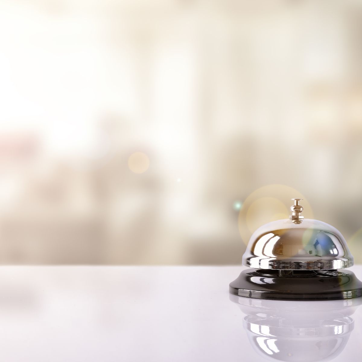 A silver service bell sitting on a white surface with a softly blurred background and warm light reflections.